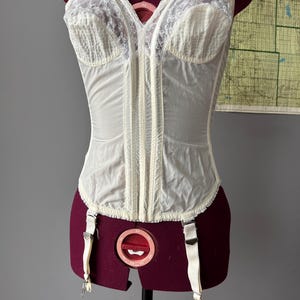 May include: Ivory-colored vintage corset with a front zipper closure and attached garter straps. The corset features delicate lace detailing on the bust and vertical ribbing throughout the bodice. It is displayed on a maroon dress form.