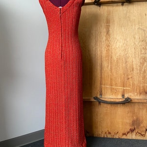 May include: A long, sleeveless, red dress with a V-neckline and a zipper down the back. The dress is made of a textured fabric with a subtle shimmer.