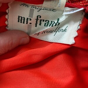 May include: A white tag with green text that reads "An original mr. frank of New York". The tag is attached to a red fabric garment.