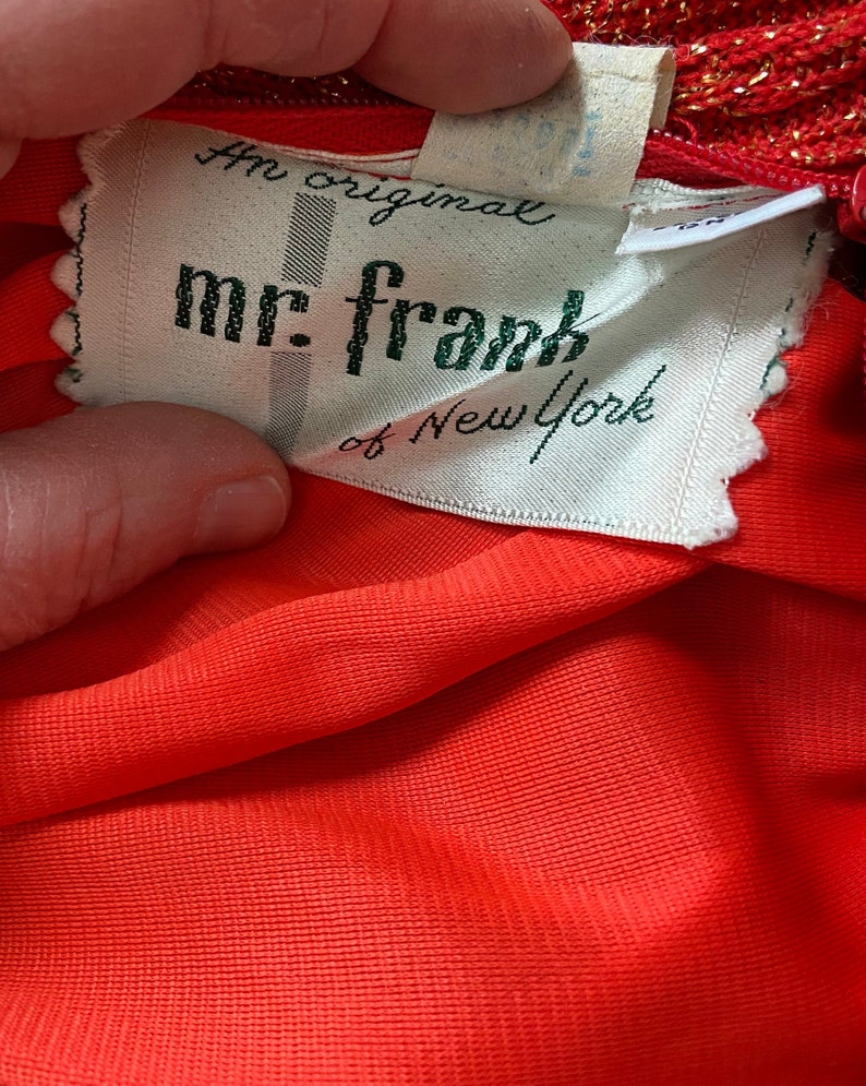May include: A white tag with green text that reads "An original mr. frank of New York". The tag is attached to a red fabric garment.