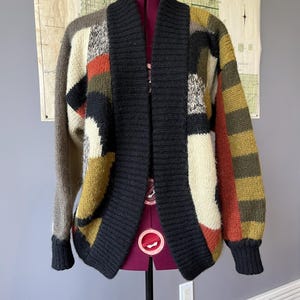 Op de afbeelding: Een veelkleurige patchwork cardigan trui met een sjaalkraag, weergegeven op een paspop. De trui heeft een mix van zwarte, olijfgroene, beige en roestkleurige panelen. De mouwen en het lichaam zijn gemaakt van verschillende geometrische vormen.