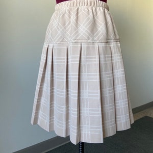 May include: A beige pleated skirt with a white checked pattern. The skirt has an elastic waistband.