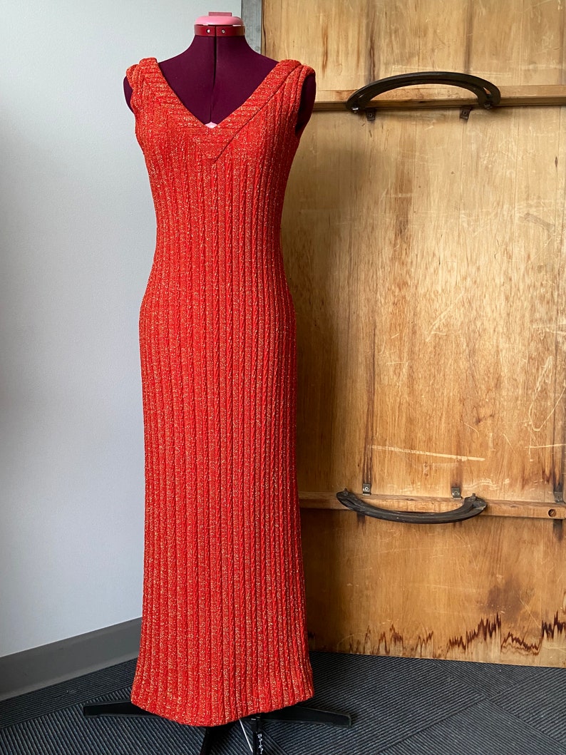 May include: A long, sleeveless, orange dress with a V-neckline and a subtle shimmer. The dress is made of a textured fabric and has a fitted silhouette.