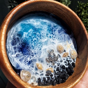 Könnte beinhalten: Eine Holzschale mit einer Harz-Ozeanszene. Die Schale zeigt eine detaillierte Darstellung von Meereswellen in Blau- und Weißtönen, mit einer kleinen silbernen Schildkröte, Muscheln und schwarzen Felsen. Die Schale wird in einer Hand gehalten.
