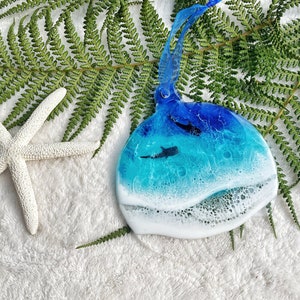Könnte beinhalten: Ein runder, blauer und weißer Harz-Anhänger mit einem weißen Wellendesign und einer Silhouette eines Hais, der im blauen Wasser schwimmt.