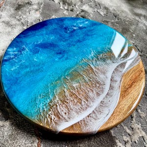 Puede incluir: Tabla redonda de madera para servir con un diseño de resina que representa una escena de playa. Presenta un océano azul vibrante, olas blancas y una orilla arenosa. El borde de la tabla es de color madera natural. Aproximadamente 30 cm de diámetro.