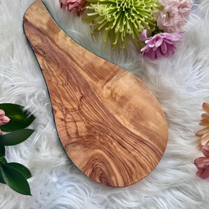 May include: A wooden serving board with a unique teardrop shape. The board features a natural wood grain pattern in shades of brown and tan. The edge is outlined with a dark blue trim. The board is surrounded by colorful flowers.