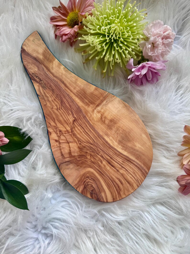 May include: A wooden serving board with a unique teardrop shape. The board features a natural wood grain pattern in shades of brown and tan. The edge is outlined with a dark blue trim. The board is surrounded by colorful flowers.