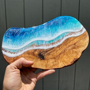 Pode incluir: Uma bandeja de servir de madeira com um design de onda de oceano de resina azul e branca. A bandeja &eacute; feita de madeira de oliveira e tem uma veia de madeira natural.