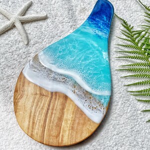 May include: A wooden serving board with a resin ocean design. The board features a light brown wooden base, transitioning to a sandy beach effect, and then to a turquoise and deep blue ocean design. A starfish is visible in the background.