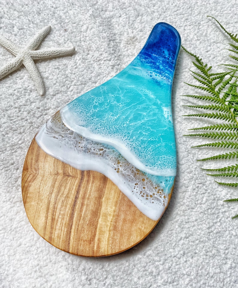 May include: A wooden serving board with a resin ocean design. The board features a light brown wooden base, transitioning to a sandy beach effect, and then to a turquoise and deep blue ocean design. A starfish is visible in the background.