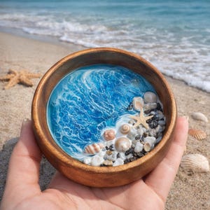 Könnte beinhalten: Eine Holzschale mit einer Miniatur-Ozeanszene. Die Schale zeigt einen blau-weißen Kunstharz-Ozean mit Muscheln, Kieselsteinen und einem Seestern. Die Schale wird in einer Hand gehalten, mit einem Sandstrand und dem Ozean im Hintergrund.