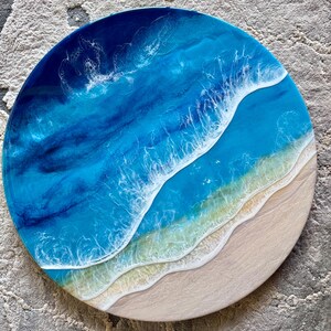 May include: Circular resin artwork showing an ocean scene. The piece uses shades of blue and white to represent the water, with a sandy beach area. The circular shape creates a coastal theme, reminiscent of the seaside.