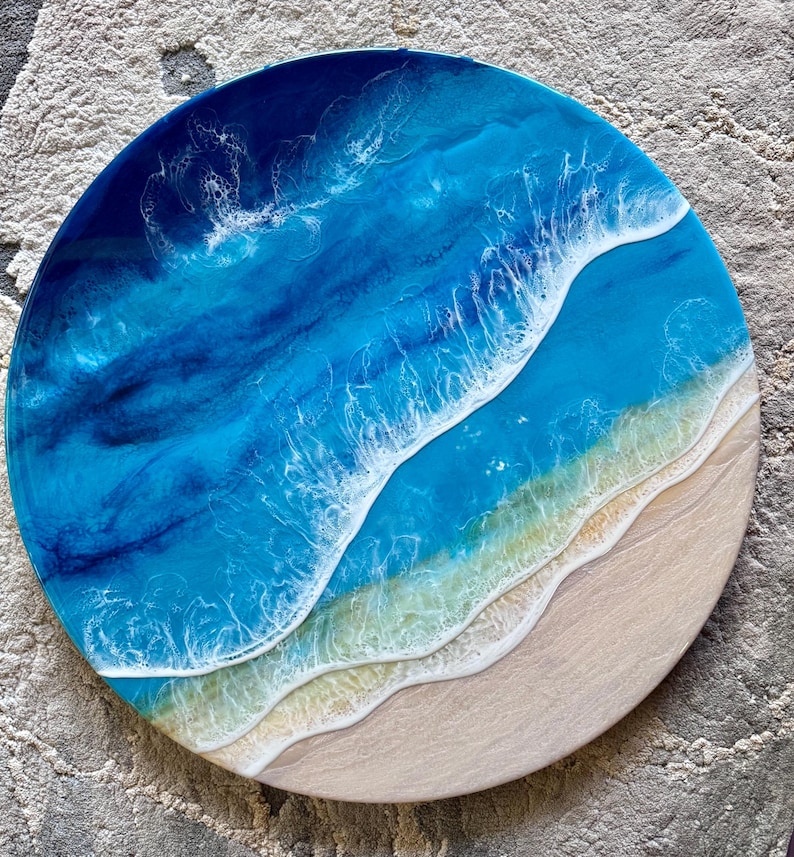 May include: Circular resin artwork showing an ocean scene. The piece uses shades of blue and white to represent the water, with a sandy beach area. The circular shape creates a coastal theme, reminiscent of the seaside.