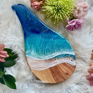 May include: A decorative wooden serving board with a resin ocean design. The board features a blue and white ocean scene with a sandy beach effect. The board is shaped like a spoon rest and is surrounded by flowers.