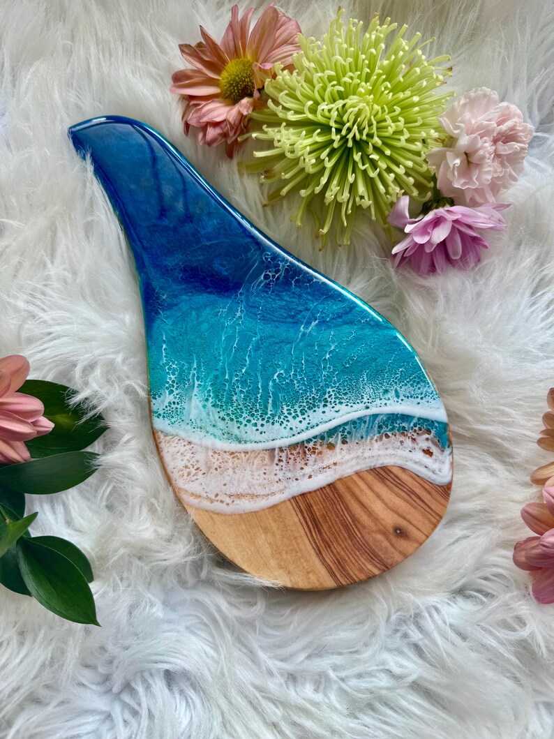 May include: A decorative wooden serving board with a resin ocean design. The board features a blue and white ocean scene with a sandy beach effect. The board is shaped like a spoon rest and is surrounded by flowers.