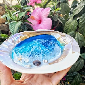 Könnte beinhalten: Eine dekorative Muschel mit einer Harz-Ozeanszene. Die Meereskunst zeigt Blau- und Weißtöne, mit einem Seestern und kleinen Muscheln. Die Muschel hat ein perlmuttartiges Finish.
