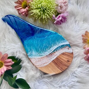 May include: A wooden spoon rest with a blue and white resin ocean design. The resin art depicts waves crashing on a sandy beach. The spoon rest is surrounded by pink and green flowers.