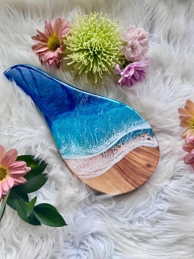 May include: A wooden spoon rest with a blue and white resin ocean design. The resin art depicts waves crashing on a sandy beach. The spoon rest is surrounded by pink and green flowers.