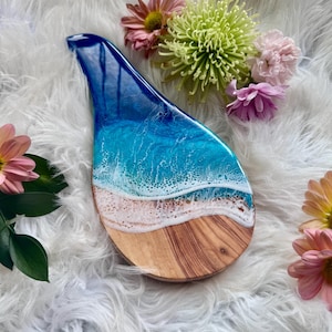 May include: A teardrop-shaped wooden serving board with a resin ocean design. The board features a blue and white ocean wave design over a sandy beach and wood grain base. Pink and yellow flowers are arranged around the board.