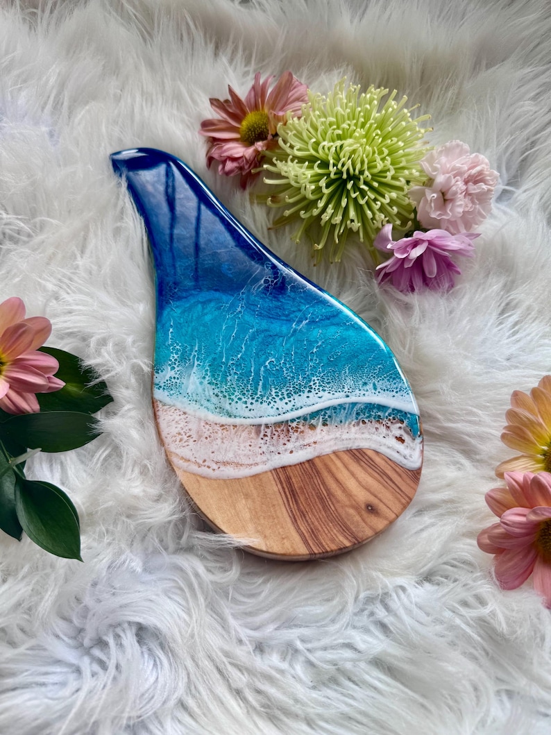 May include: A teardrop-shaped wooden serving board with a resin ocean design. The board features a blue and white ocean wave design over a sandy beach and wood grain base. Pink and yellow flowers are arranged around the board.