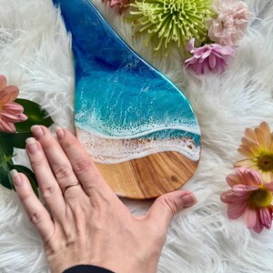 May include: A wooden serving board with a resin ocean design. The board features a blue and white wave pattern over a sandy beach design. The board is surrounded by pink and yellow flowers.