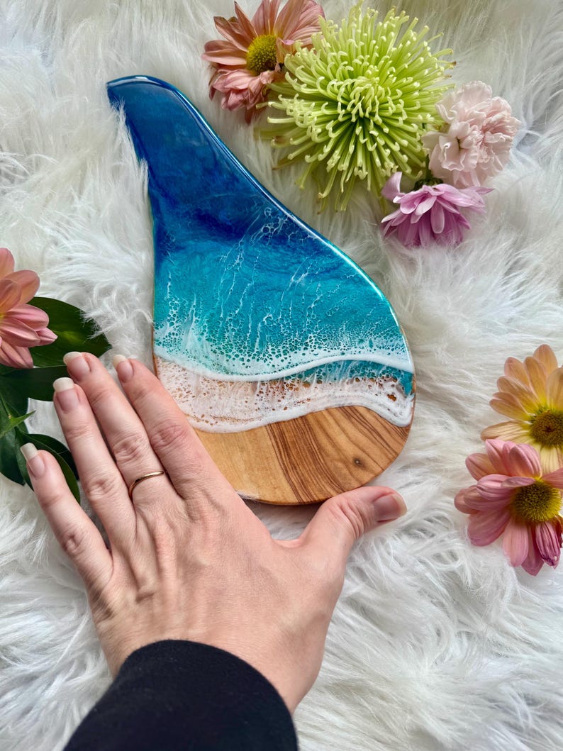 May include: A wooden serving board with a resin ocean design. The board features a blue and white wave pattern over a sandy beach design. The board is surrounded by pink and yellow flowers.