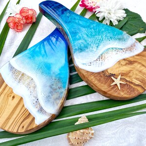 May include: Two wooden serving boards with a unique ocean wave design. The boards feature a natural wood grain base with a white and blue resin wave design. A starfish is placed on one board, with a seashell and flowers in the background.