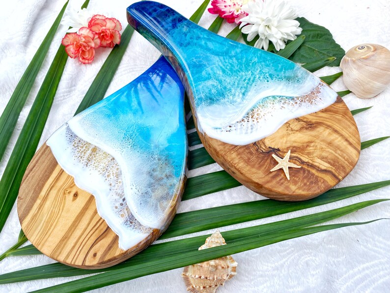 May include: Two wooden serving boards with a unique ocean wave design. The boards feature a natural wood grain base with a white and blue resin wave design. A starfish is placed on one board, with a seashell and flowers in the background.