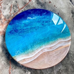 May include: Circular resin artwork showing a beach scene. It features a gradient of blues for the ocean, blending into a sandy beige shoreline. The glossy resin has wave-like patterns and a smooth finish. The diameter is approximately 25 cm.
