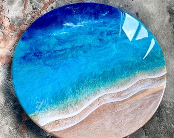 May include: Round resin art piece depicting a beach scene. The artwork features a gradient of blues representing the ocean, transitioning to a sandy beige shoreline. The resin is glossy, with visible wave-like patterns and a smooth finish.