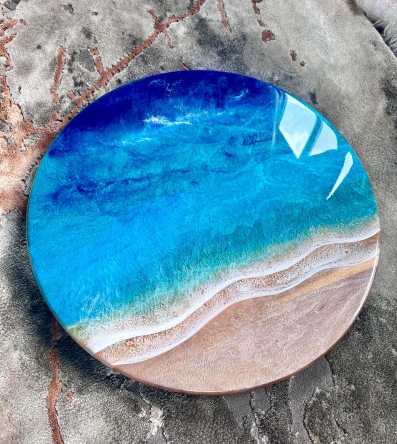 May include: Circular resin artwork showing a beach scene. It features a gradient of blues for the ocean, blending into a sandy beige shoreline. The glossy resin has wave-like patterns and a smooth finish. The diameter is approximately 25 cm.