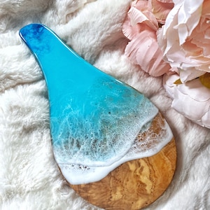 May include: A decorative wooden serving board with a resin design. The top portion features a vibrant blue resin that transitions to white, mimicking ocean waves. The bottom is natural wood, creating a beach-themed aesthetic.