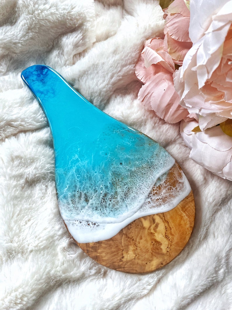 May include: A decorative wooden serving board with a resin design. The top portion features a vibrant blue resin that transitions to white, mimicking ocean waves. The bottom is natural wood, creating a beach-themed aesthetic.