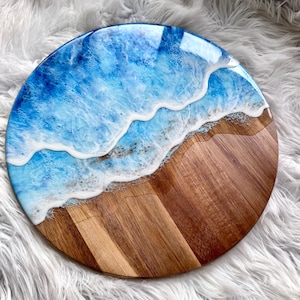 May include: A round wooden serving board featuring a glossy resin art design that mimics an ocean shoreline. The board displays a natural brown wood grain on one side and a vibrant blue and white wave pattern on the other side.