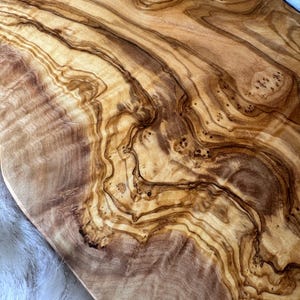 Pode incluir: Uma t&aacute;bua de servir de madeira de oliveira grande e oval com um acabamento natural de cor castanho claro. A veia da madeira &eacute; vis&iacute;vel e tem um padr&atilde;o &uacute;nico e em espiral. A t&aacute;bua &eacute; perfeita para servir queijo, charcutaria ou outros aperitivos.
