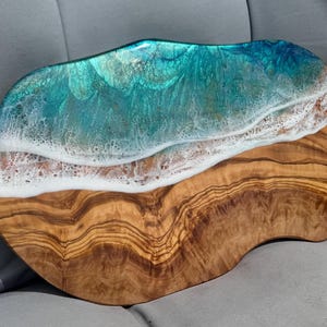 Pode incluir: Uma t&aacute;bua de servir de madeira com um design de resina ep&oacute;xi azul e branco que se assemelha a ondas do oceano. A t&aacute;bua tem um padr&atilde;o de gr&atilde;o de madeira natural e uma forma &uacute;nica e irregular.