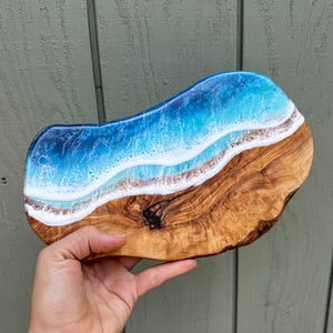 Pode incluir: Uma t&aacute;bua de servir de madeira com um design de onda de oceano de resina azul e branca. A t&aacute;bua &eacute; feita de madeira de oliveira e tem um acabamento natural e r&uacute;stico.