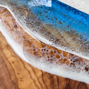 Pode incluir: Uma t&aacute;bua de servir de madeira com um design de resina ep&oacute;xi azul e branca que se assemelha a uma onda do oceano. A resina tem uma textura bolhosa e a madeira tem um padr&atilde;o de gr&atilde;o natural.