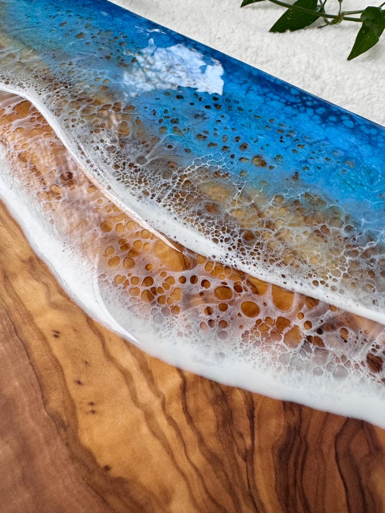 Pode incluir: Uma t&aacute;bua de servir de madeira com um design de resina ep&oacute;xi azul e branca que se assemelha a uma onda do oceano. A resina tem uma textura bolhosa e a madeira tem um padr&atilde;o de gr&atilde;o natural.