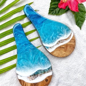 May include: Two wooden serving spoons with a unique ocean wave design. The spoons feature a light brown wooden base with a white and blue resin top, mimicking ocean waves. A red hibiscus flower and green leaves are in the background.