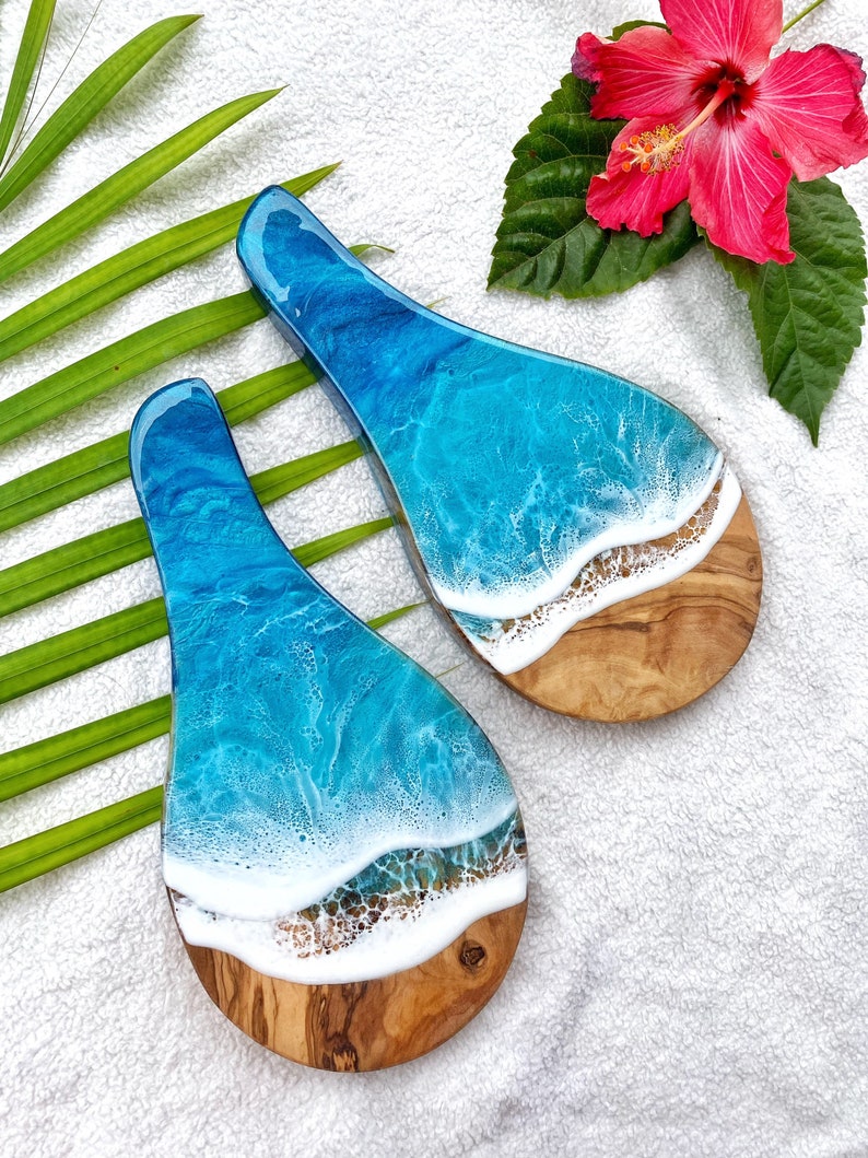 May include: Two wooden serving spoons with a unique ocean wave design. The spoons feature a light brown wooden base with a white and blue resin top, mimicking ocean waves. A red hibiscus flower and green leaves are in the background.