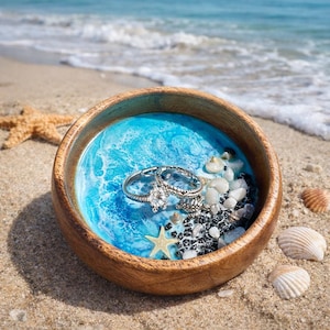 May include: A wooden bowl with a blue resin ocean design, holding several silver rings with clear gemstones. The bowl is decorated with small seashells, black pebbles, and a starfish. The scene is set on a sandy beach with the ocean in the background.