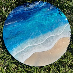 May include: A circular resin artwork showing an ocean scene. The piece uses shades of blue and turquoise to represent the water, with white foamy waves and a sandy beach. The artwork is set against a backdrop of green grass.