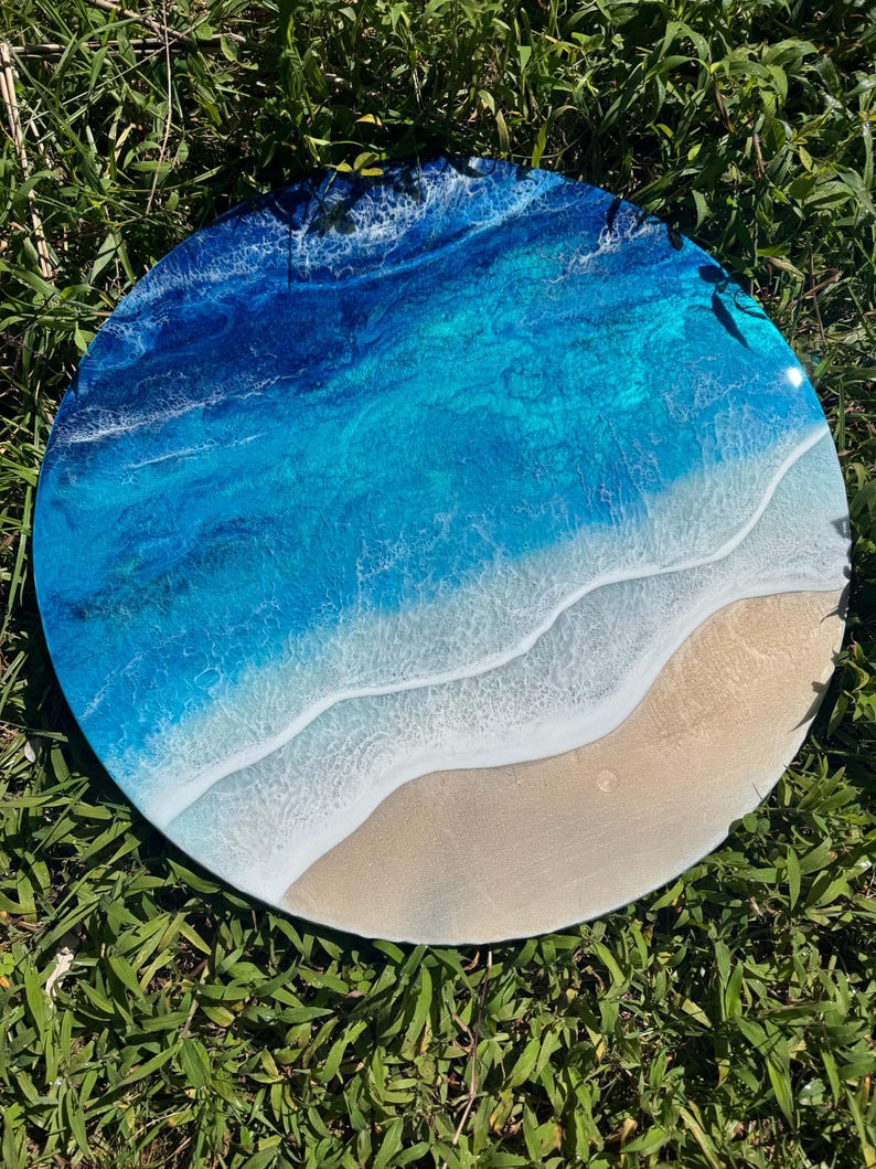 May include: A circular resin artwork showing an ocean scene. The piece uses shades of blue and turquoise to represent the water, with white foamy waves and a sandy beach. The artwork is set against a backdrop of green grass.