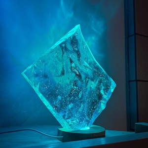 May include: A blue resin lamp with a unique, organic shape. The lamp is lit from within, casting a soft blue glow. It sits on a wooden base.