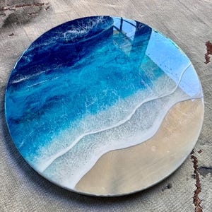 May include: Circular resin artwork showing an ocean scene. The piece uses shades of blue and white to represent the water, with a sandy beach area. The diameter of the artwork is approximately 30 cm.