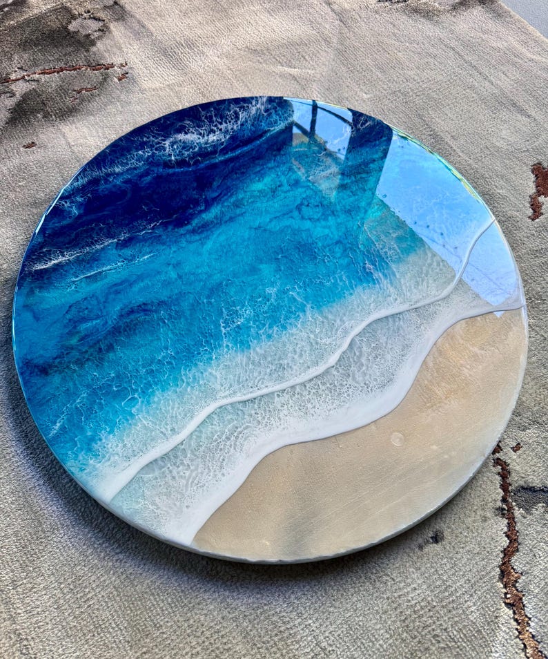 May include: Circular resin artwork showing an ocean scene. The piece uses shades of blue and white to represent the water, with a sandy beach area. The diameter of the artwork is approximately 30 cm.