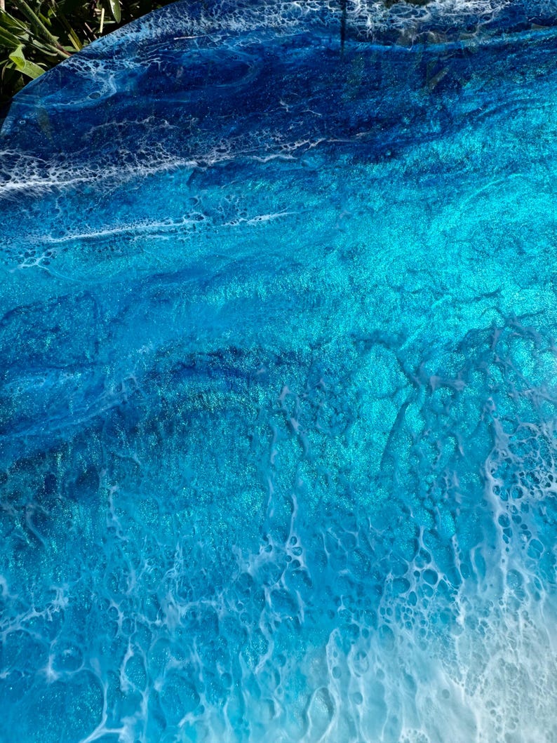 May include: Abstract artwork with a coastal theme. The image shows a close-up of a surface with layers of blue and turquoise resin, resembling ocean waves. The resin has a glossy finish with white foamy details.