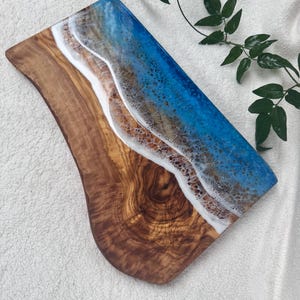 Pode incluir: Uma t&aacute;bua de servir de madeira com um design de resina ep&oacute;xi azul e branco que se assemelha a ondas do oceano. A t&aacute;bua tem uma veia de madeira natural e uma borda curva.